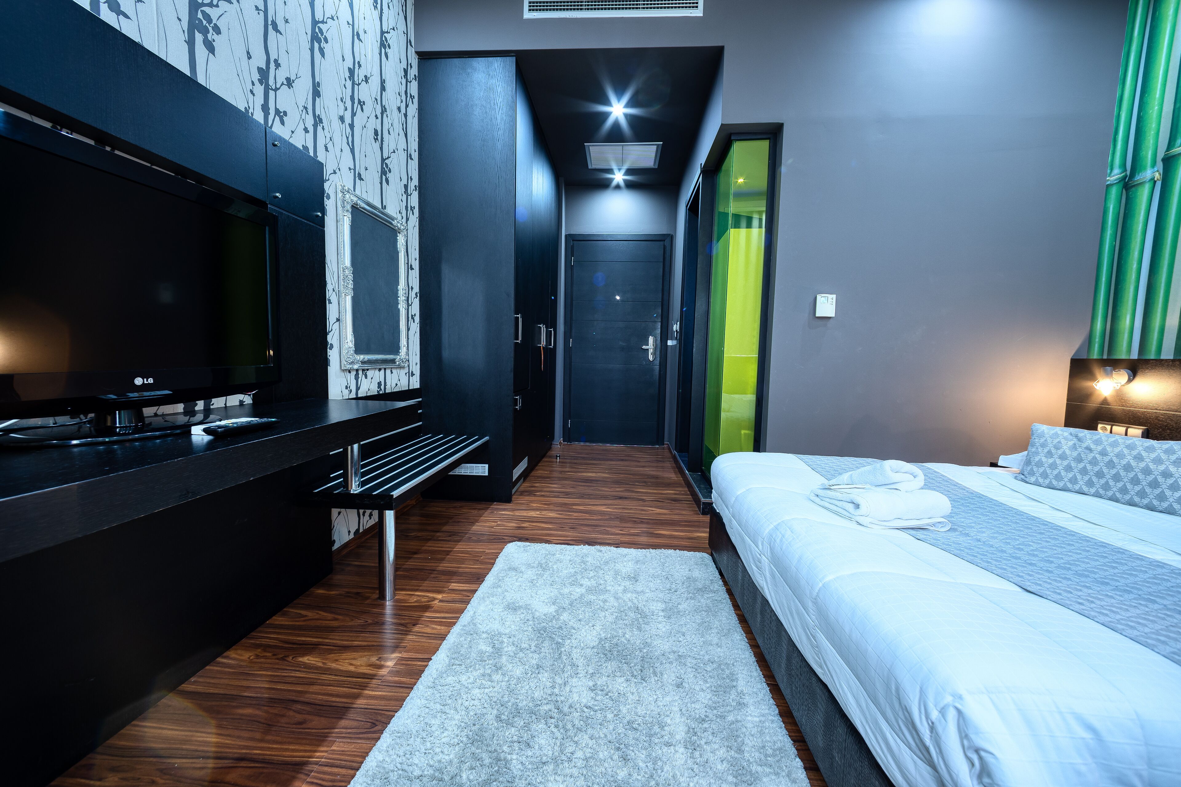 A modern hotel room with a flat-screen TV, a bed with white linens, a grey rug, and a wooden floor.