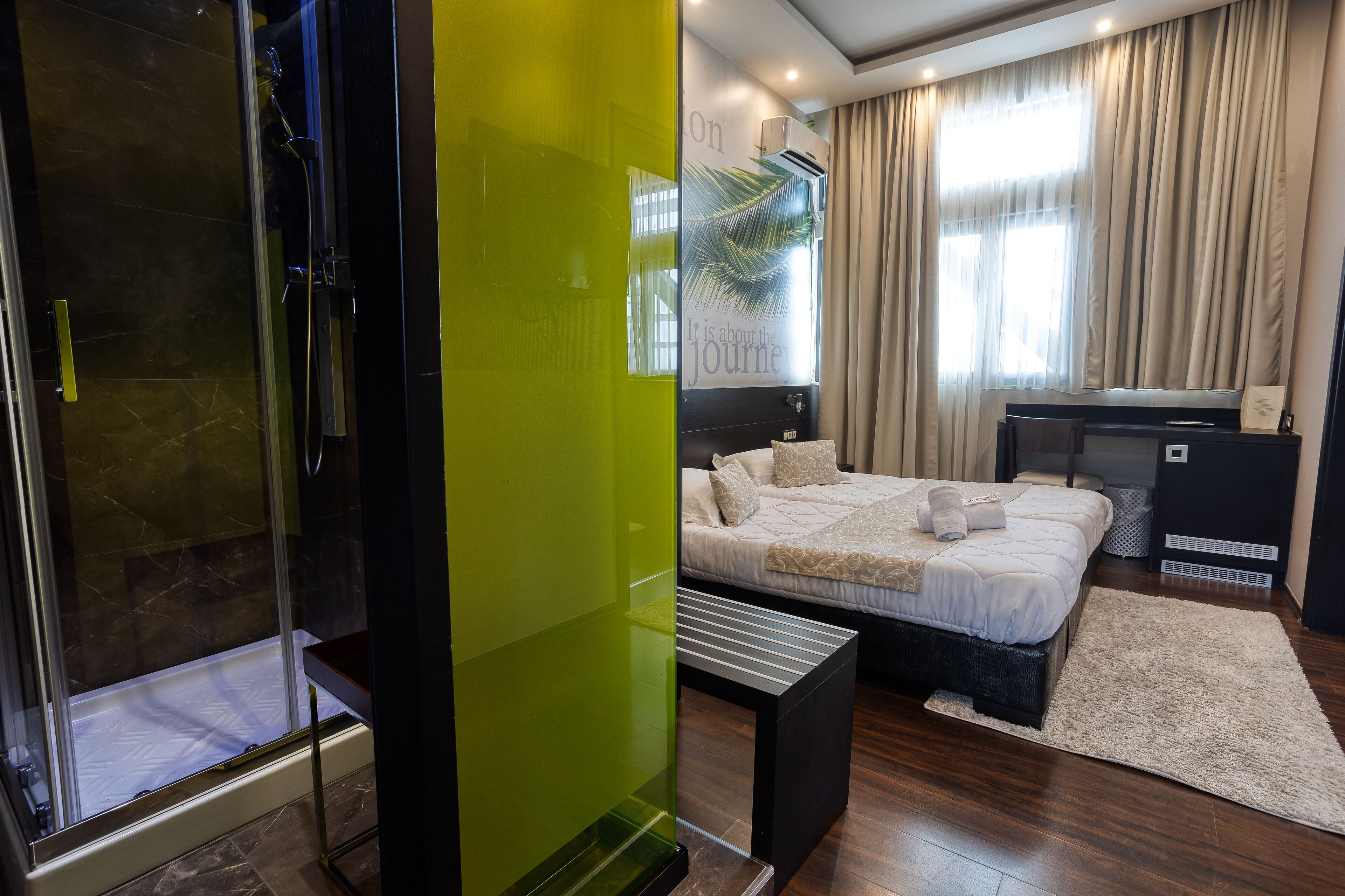A modern hotel room with a glass shower enclosure, a bed with a white comforter, a desk, and a large window with curtains.