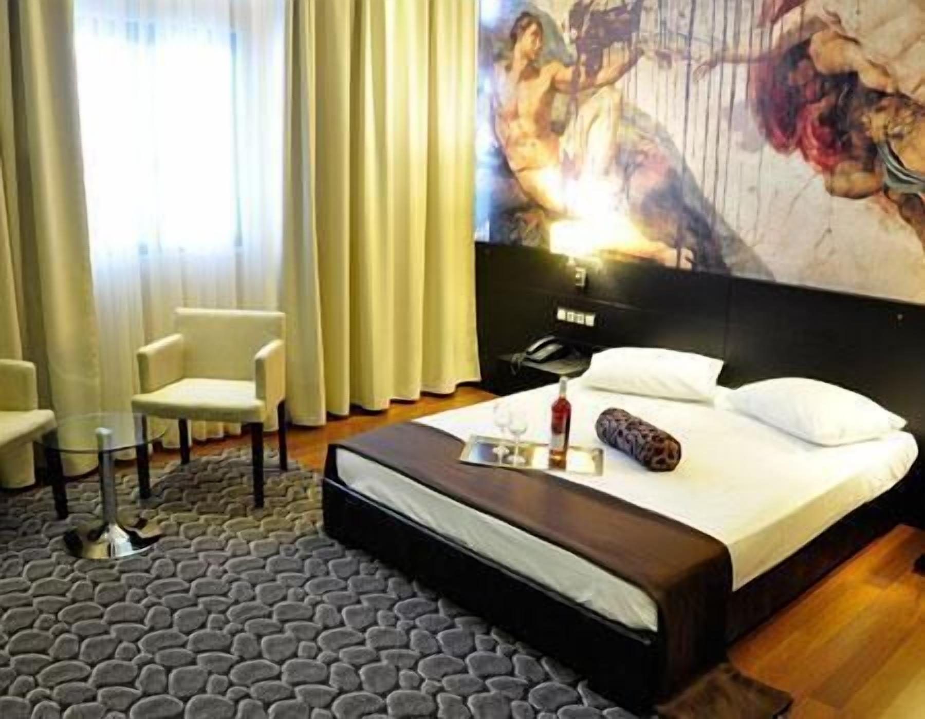 A hotel room with a bed, a bedside table, a chair, and a large painting on the wall.