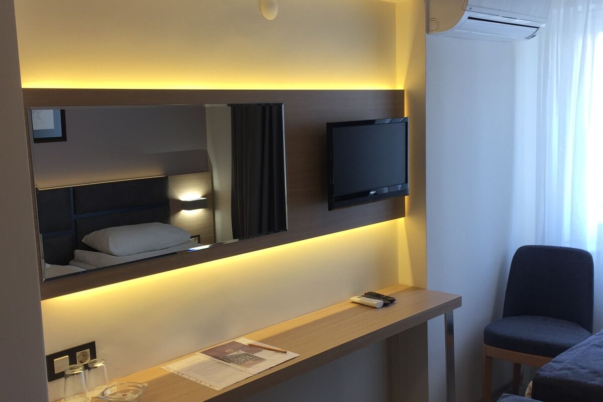 A hotel room with a bed, a desk with a chair, a TV, and an air conditioning unit.