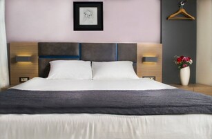 Una cama bien hecha con un edredón gris, un cabecero con detalles azules, lámparas de noche, un cuadro en la pared y un jarrón con flores en la mesita de noche.