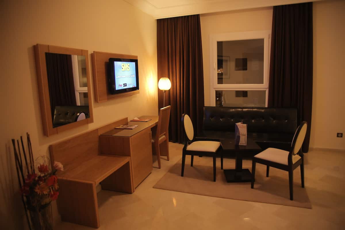 A hotel room with a wooden desk, two chairs, a sofa, a TV mounted on the wall, and a window with curtains.