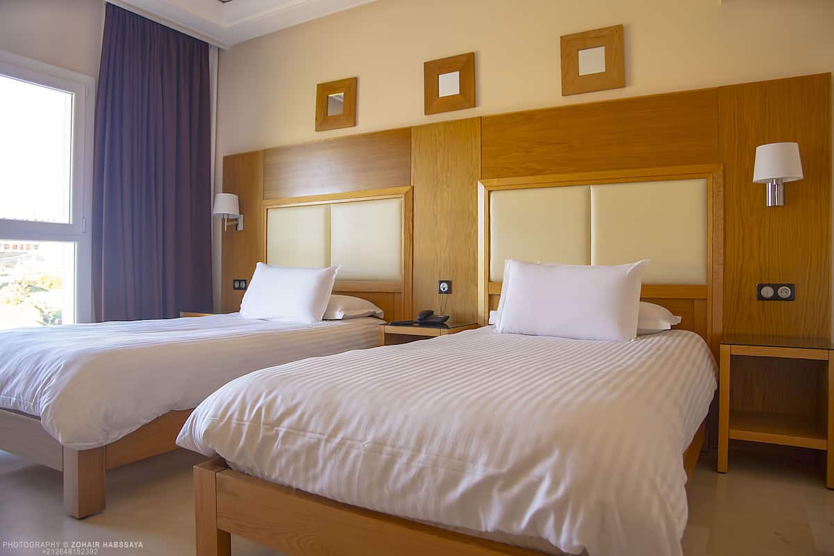 A hotel room with two beds, wooden headboards, bedside lamps, and a window with curtains.
