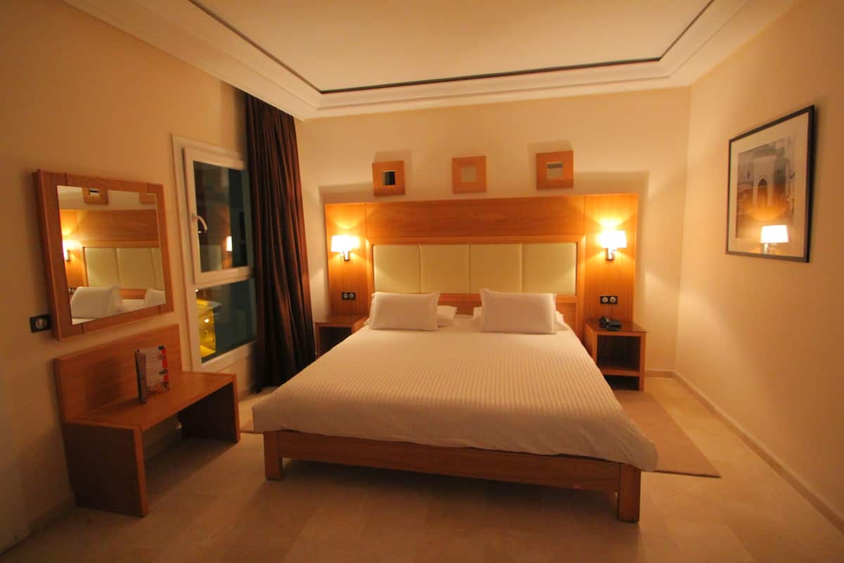 A hotel room with a large bed, bedside lamps, a wooden bench, a mirror, and a framed picture on the wall.
