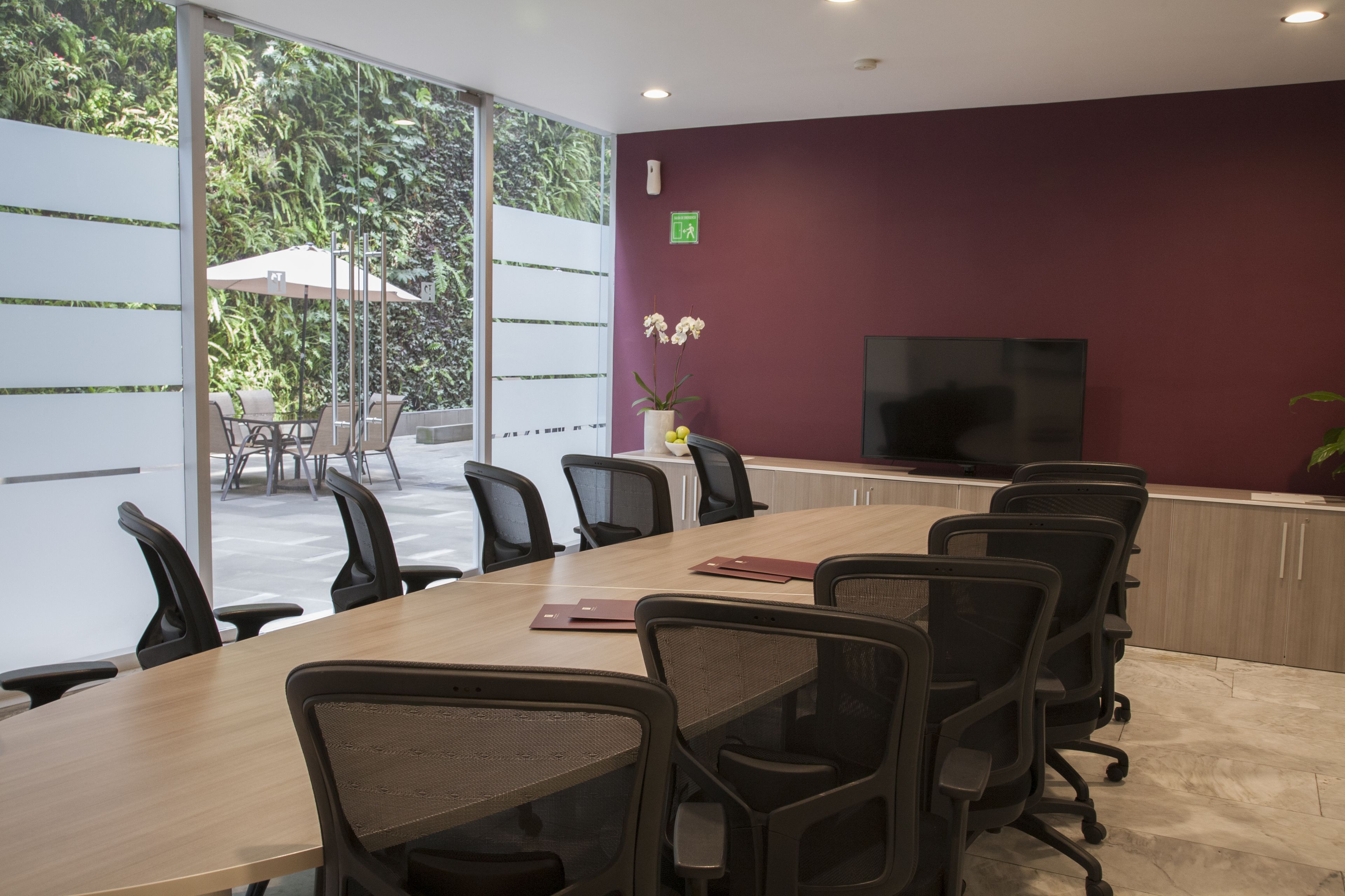 Uma sala de conferências com uma mesa comprida, várias cadeiras, uma televisão e vista para o exterior com áreas verdes.