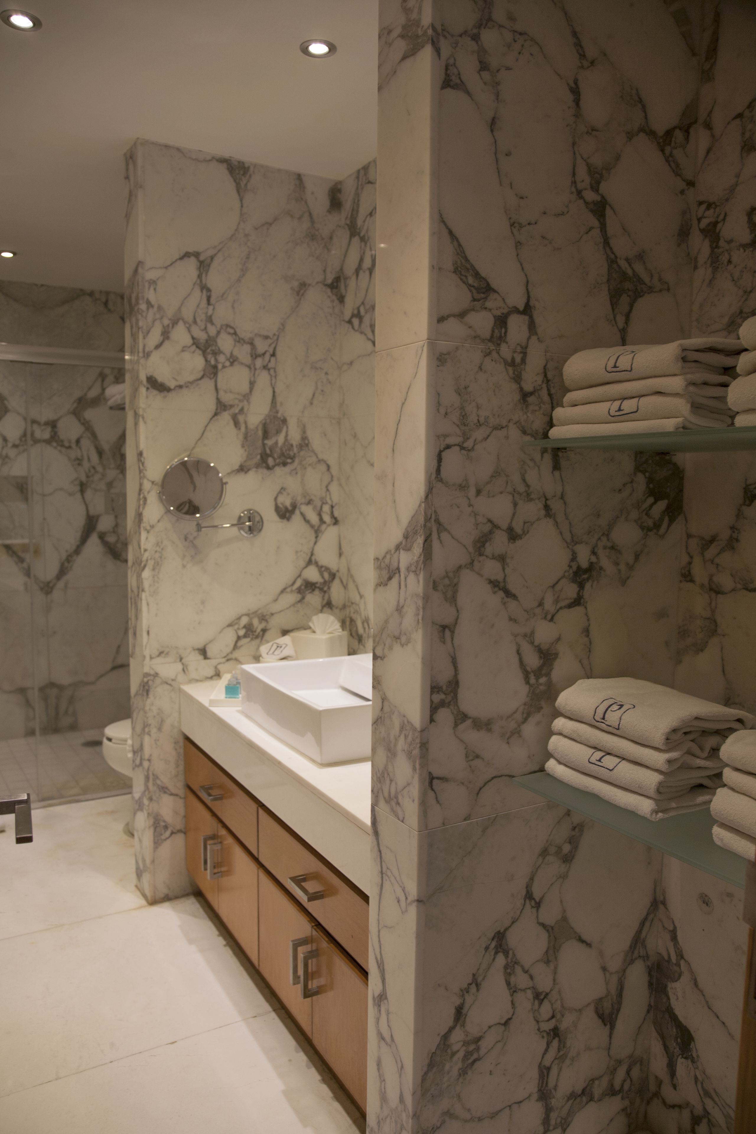 Un baño con paredes de mármol, cabina de ducha de vidrio, lavamanos con espejo y toallas ordenadas en un estante.