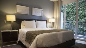 Una habitación de hotel moderna con una cama grande, dos lámparas de noche, una mesita de noche y vistas a zonas verdes a través de amplias ventanas.
