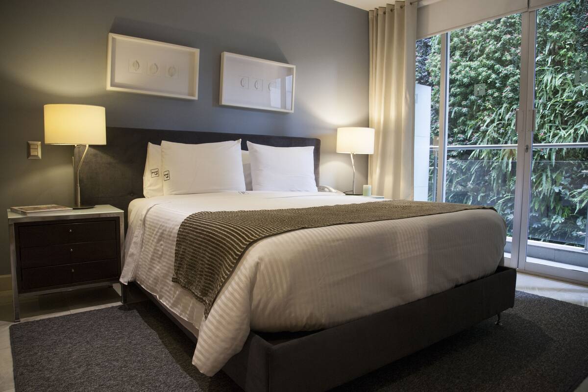 Una habitación de hotel moderna con una cama grande, dos lámparas de noche, una mesita de noche y vistas a zonas verdes a través de amplias ventanas.