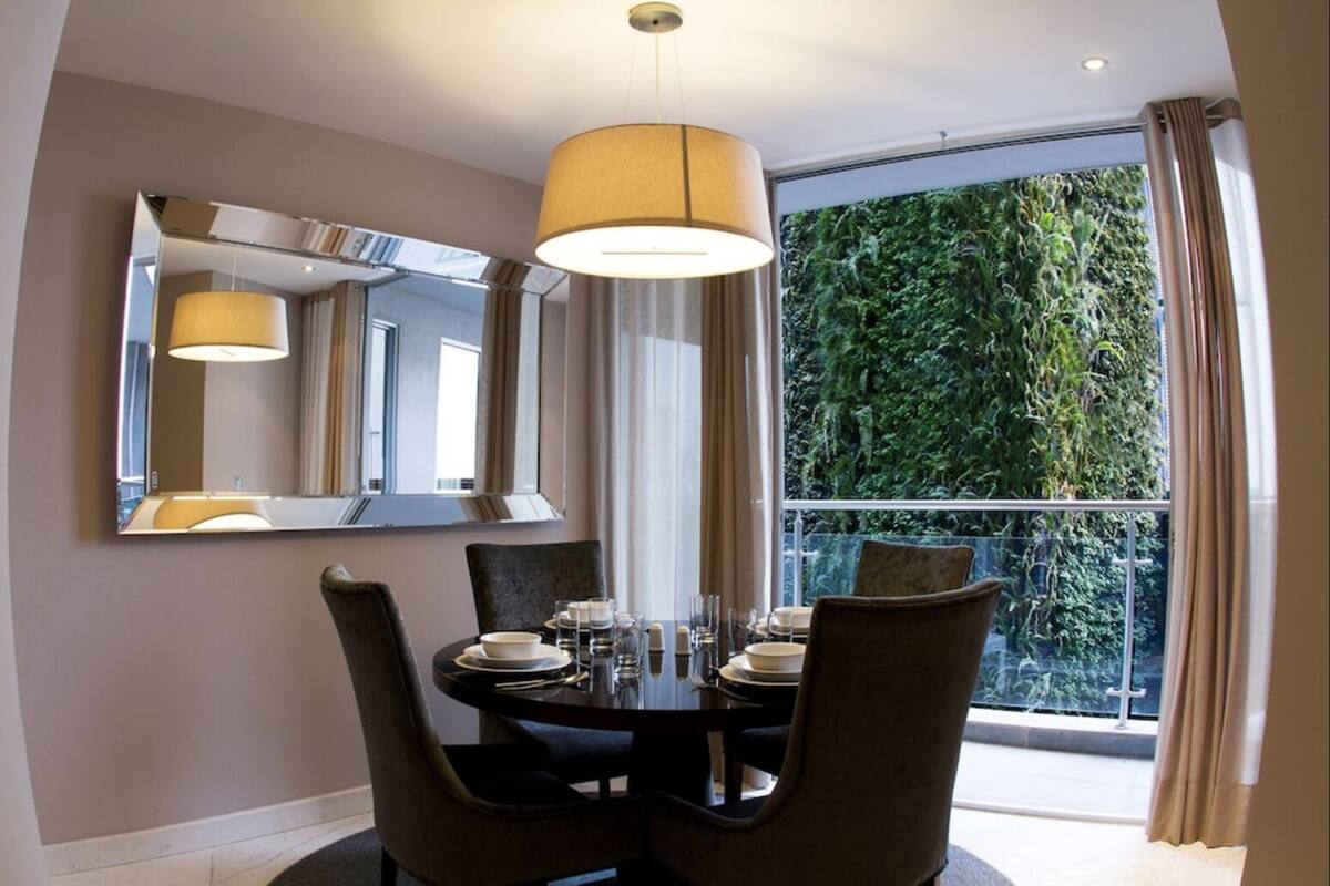 Comedor con una mesa redonda dispuesta para cuatro personas, un espejo grande y vista a zonas verdes a través de un balcón.