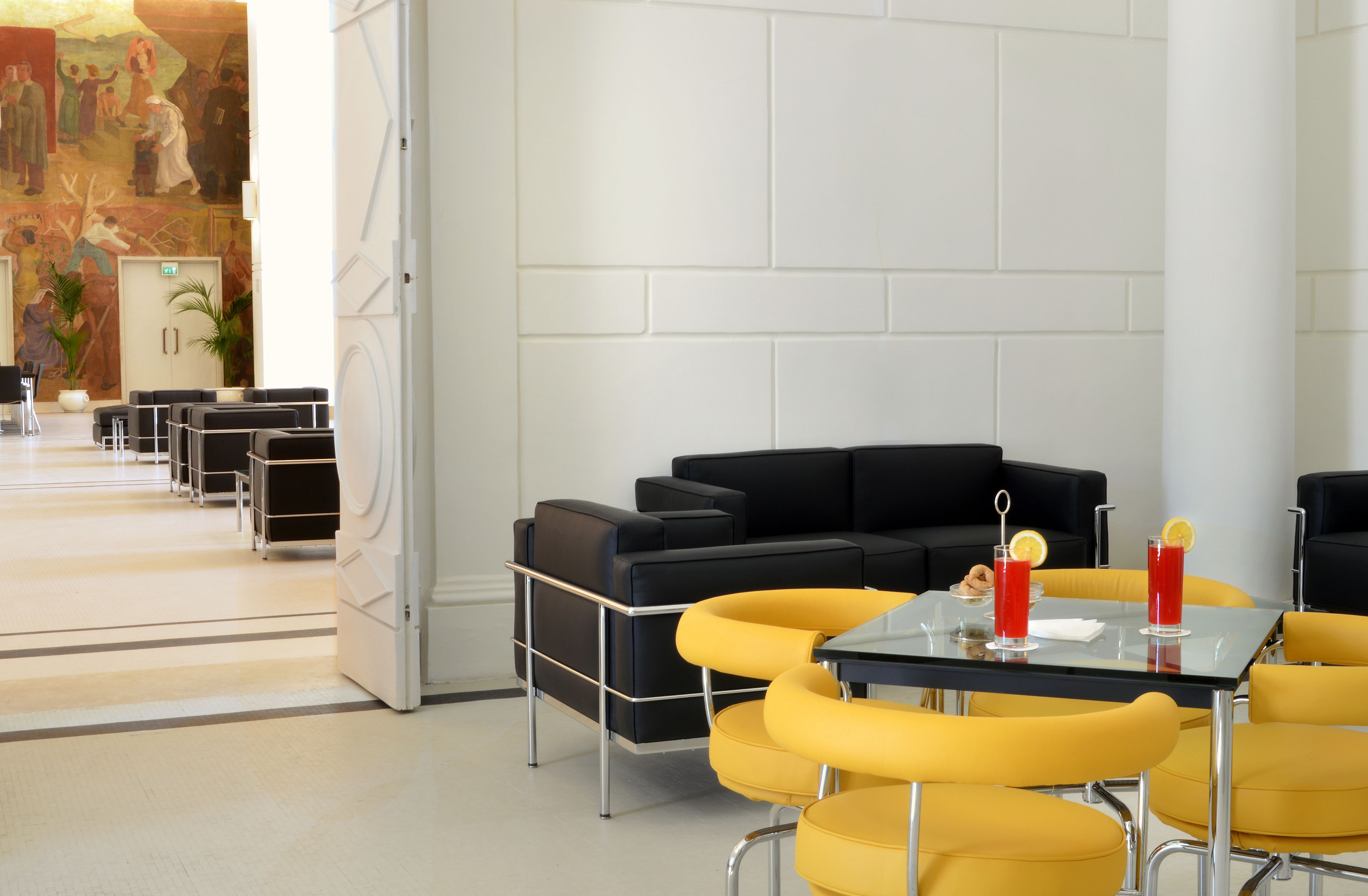 Un hall d’hôtel moderne avec un canapé en cuir noir, des fauteuils jaunes, une table en verre avec des boissons et une fresque murale.