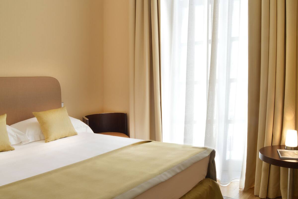 Une chambre d’hôtel avec un grand lit, une table de chevet avec une lampe et une vue dégagée à travers des rideaux légers.