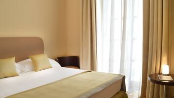 Quarto de hotel com uma cama grande, uma mesa de cabeceira com abajur e vista para o exterior através de cortinas leves.