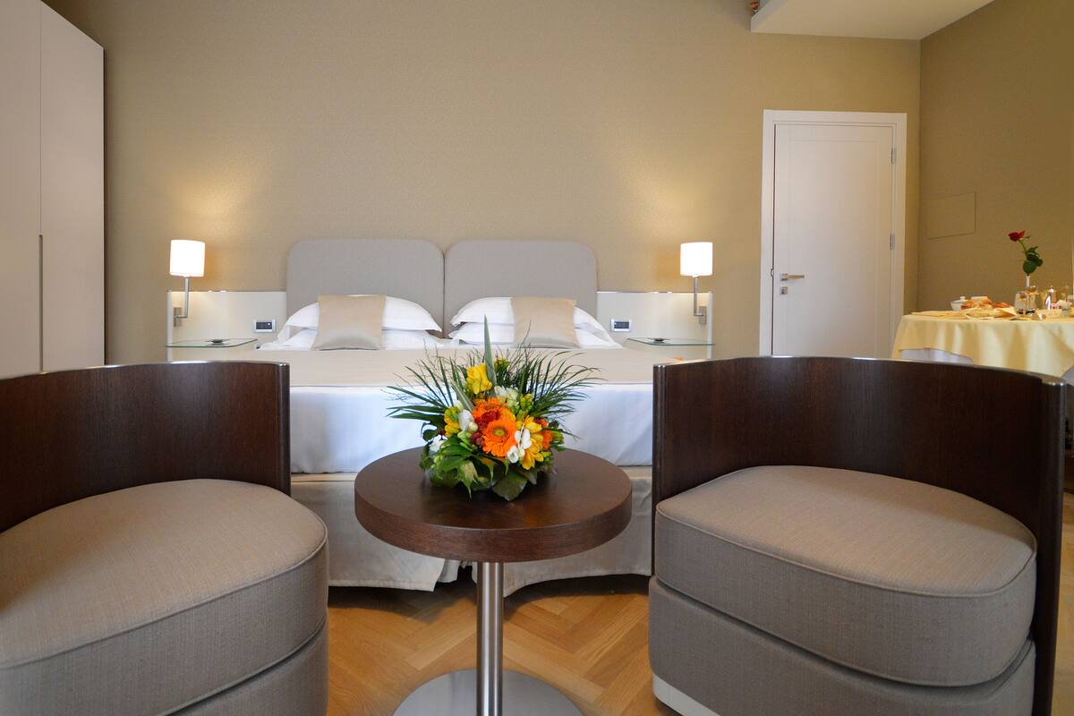 Une chambre d’hôtel comprenant un lit, deux fauteuils, une petite table ronde agrémentée d’un arrangement floral et une table à manger décorée d’un vase de fleurs.