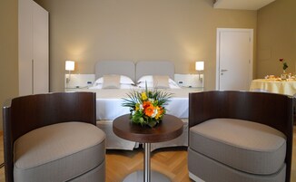 Hotelkamer met een bed, twee fauteuils, een kleine ronde tafel met bloemen en een eettafel gedekt met een bloevas.