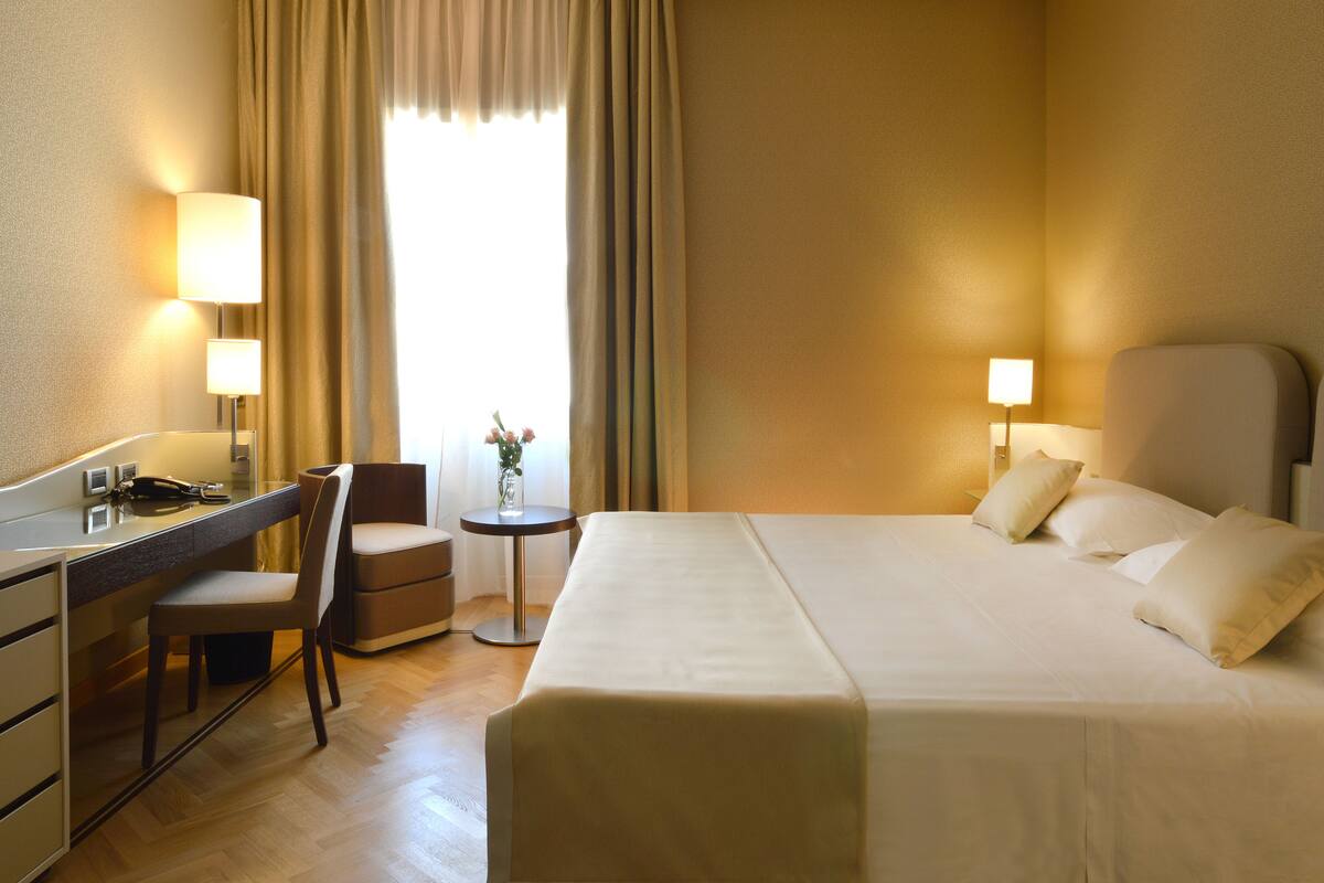 Une chambre d’hôtel avec un lit, un bureau, une chaise, une petite table ornée d’un vase de fleurs et une fenêtre avec des rideaux.
