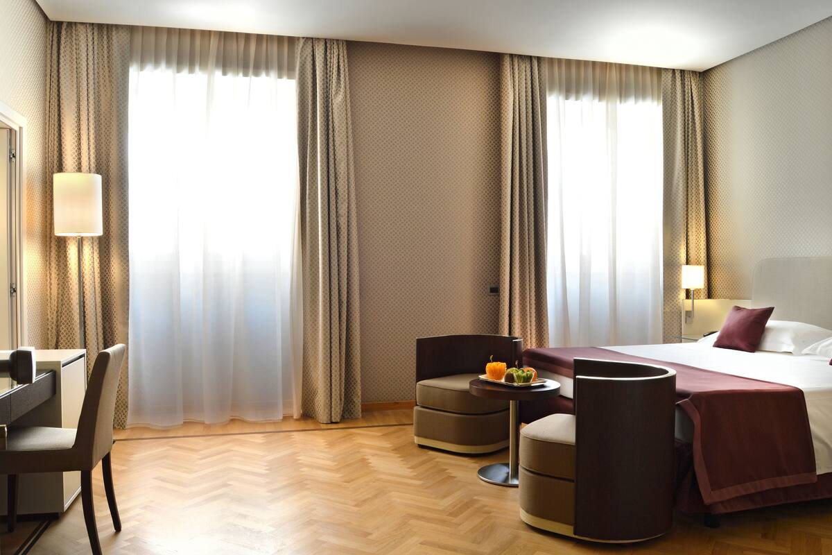 Une chambre d’hôtel avec un lit, un bureau, une chaise, une petite table avec une lampe et des rideaux.