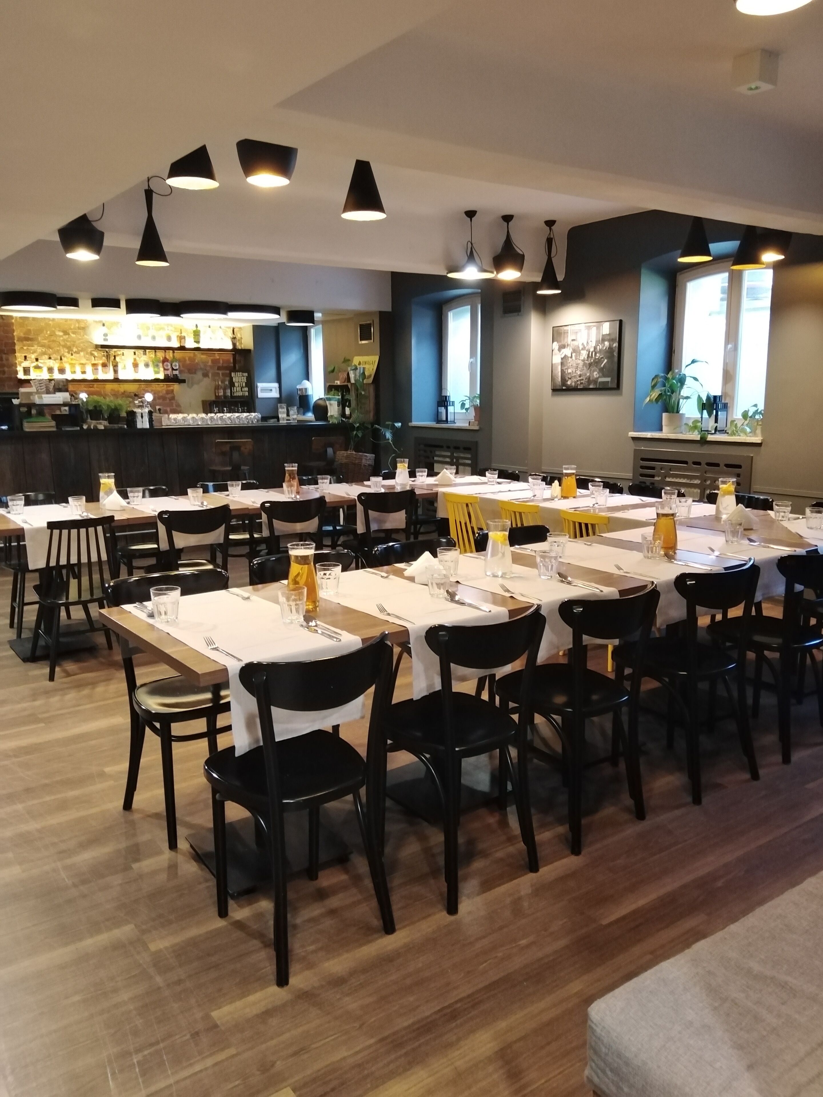 Restaurante com várias mesas e cadeiras, área de bar ao fundo e iluminação pendente.