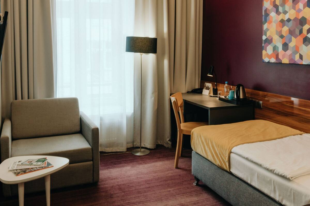 Habitación de hotel con cama, escritorio y silla, sofá, lámpara de pie y un cuadro en la pared.