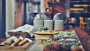 A buffet setup with various food items, including bread, salads, and condiments, in a modern dining area with brick walls and a visible kitchen in the background. - Hotel Tobaco (Lodz)