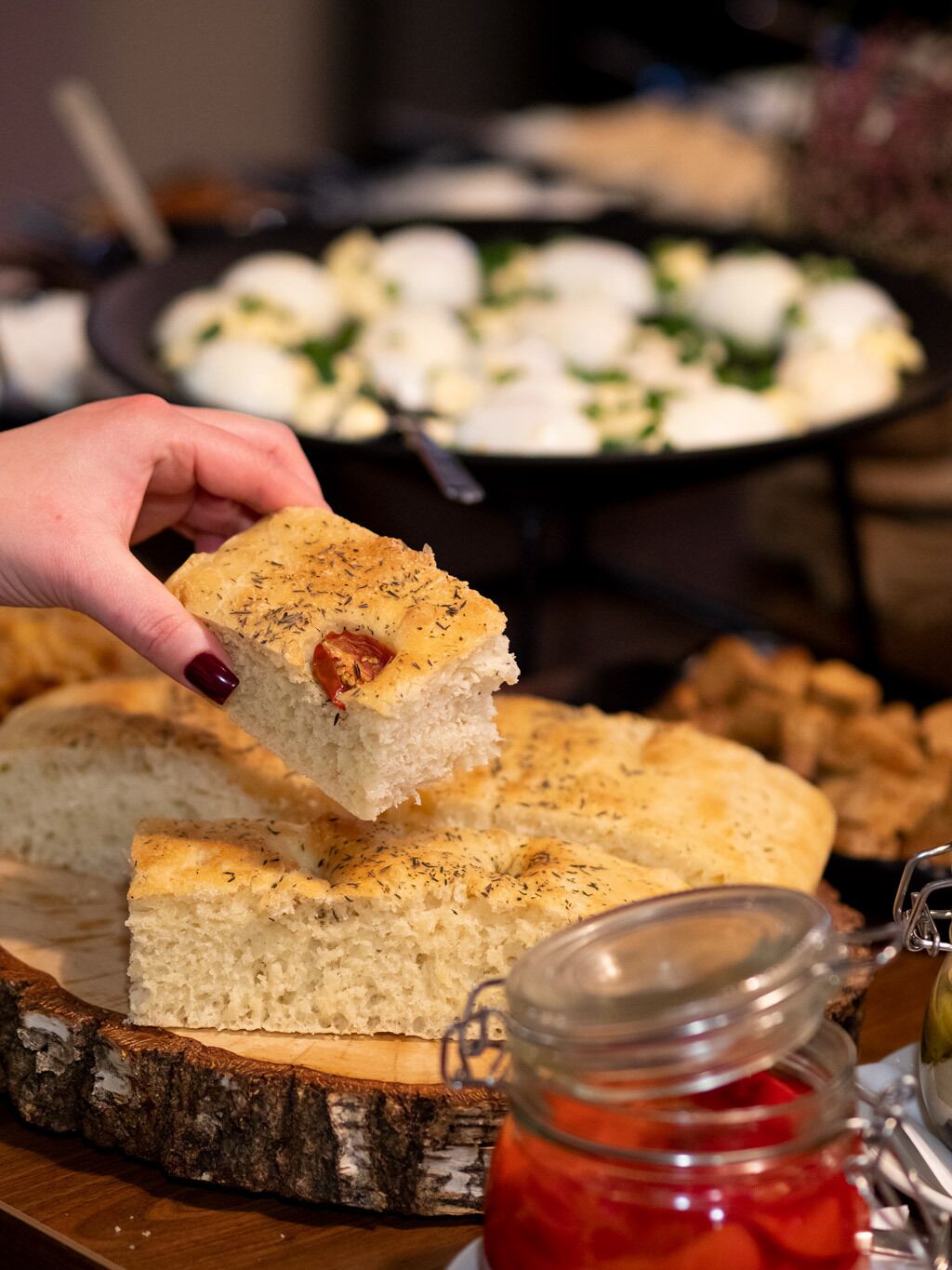 Una mano che tiene un pezzo di pane con erbe aromatiche, un barattolo di marmellata rossa e un piatto di cibo sullo sfondo.