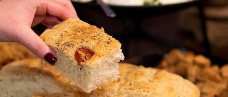Una mano che tiene un pezzo di pane con erbe aromatiche, un barattolo di marmellata rossa e un piatto di cibo sullo sfondo.