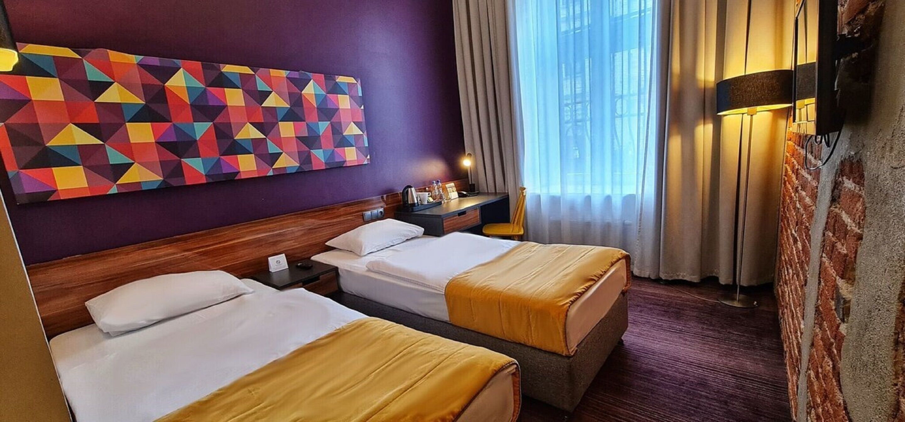 A hotel room with two beds, a colorful geometric wall art, a nightstand with a lamp, and a view of the outdoors through large windows.