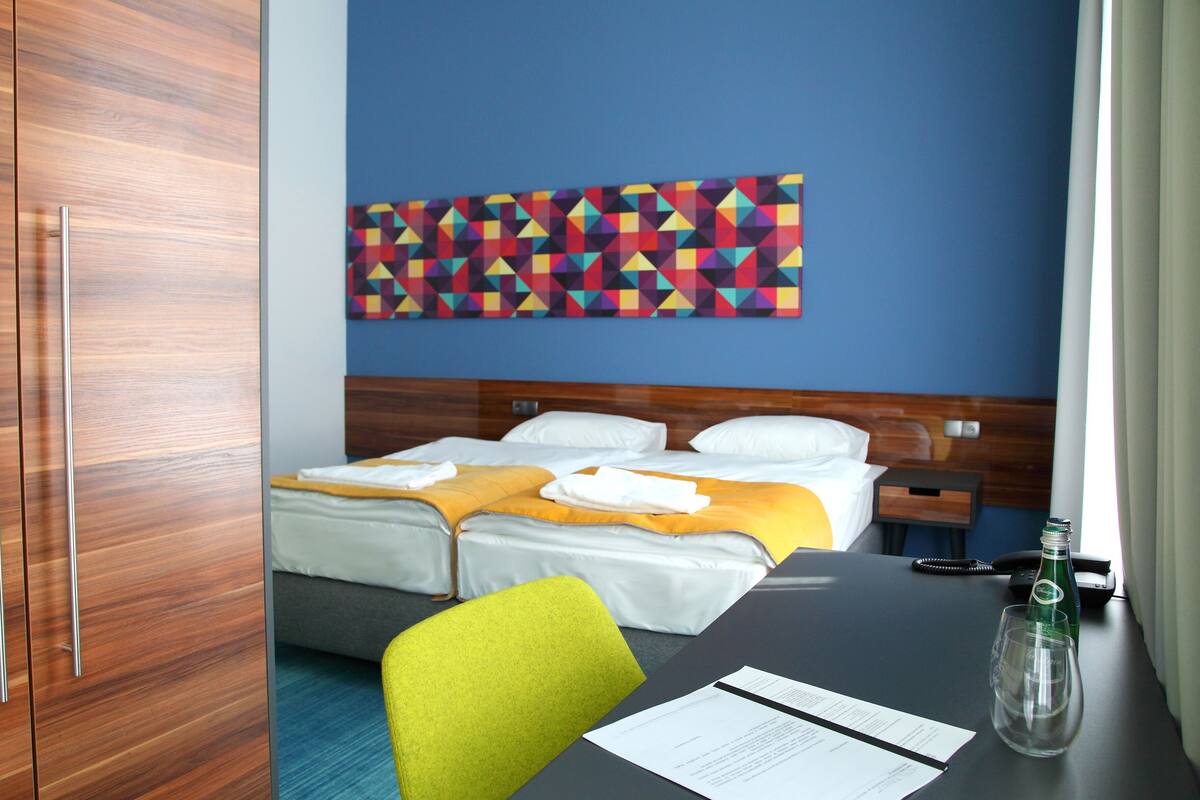 Habitación de hotel con dos camas, un armario de madera, un cuadro de pared con arte geométrico de colores, un escritorio con una botella y un vaso, y una silla.