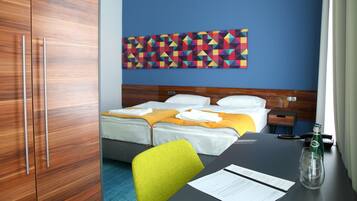 Habitación de hotel con dos camas, un armario de madera, un cuadro de pared con arte geométrico de colores, un escritorio con una botella y un vaso, y una silla.