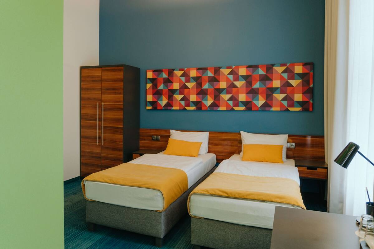 Habitación de hotel con dos camas, un armario de madera, un cuadro de arte geométrico de colores y un escritorio con lámpara.
