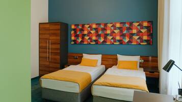 A hotel room with two beds, a wooden wardrobe, a colorful geometric wall art, and a desk with a lamp.