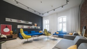 A modern living room with a sectional sofa, a yellow armchair, and a grey sofa. There are white shelves with books and decorative items, and a large window with curtains. - Hotel Tobaco (Lodz)