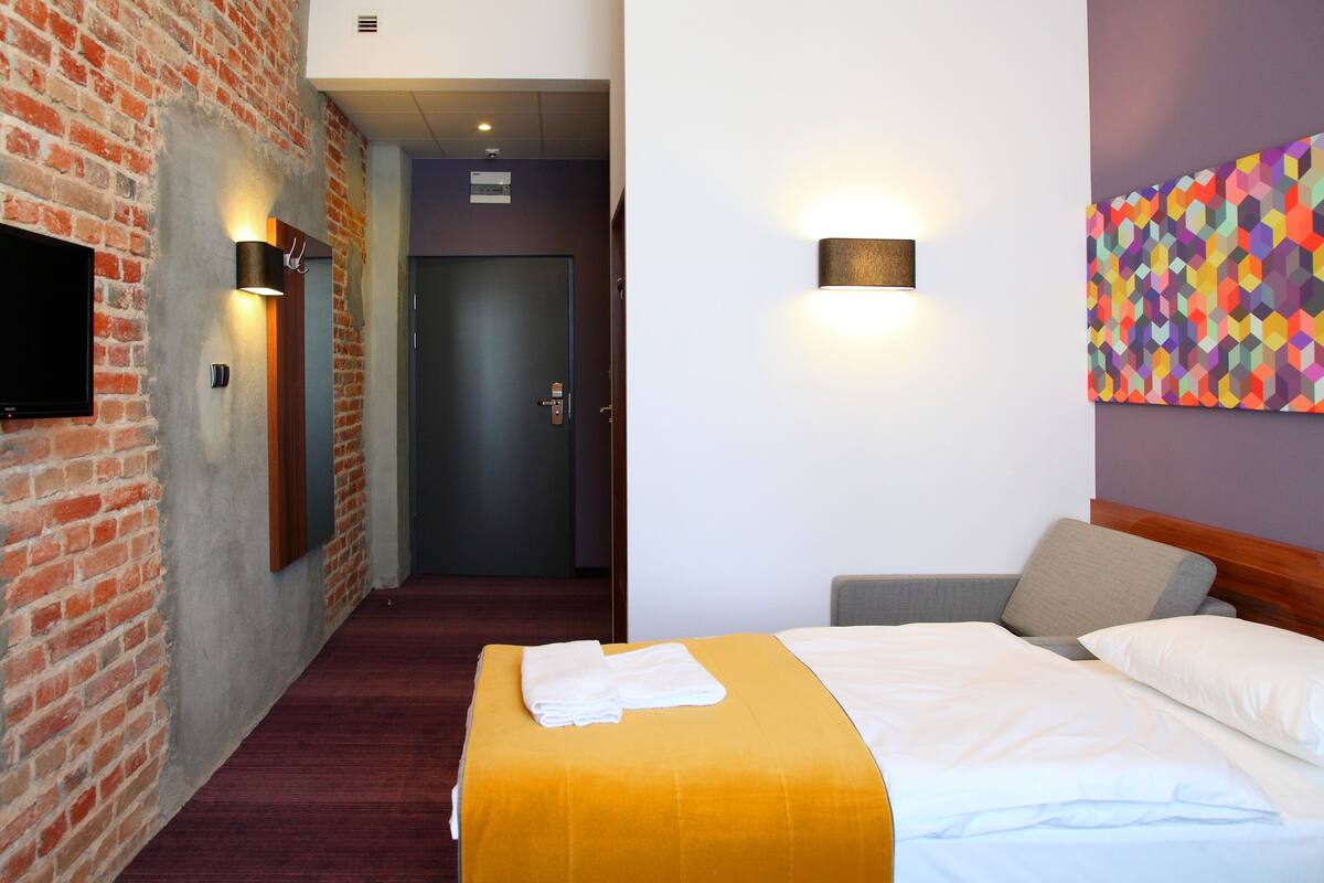Una habitación de hotel con una pared de ladrillo, un televisor de pantalla plana, una cama con cubrecama amarillo y un cuadro abstracto colorido en la pared.