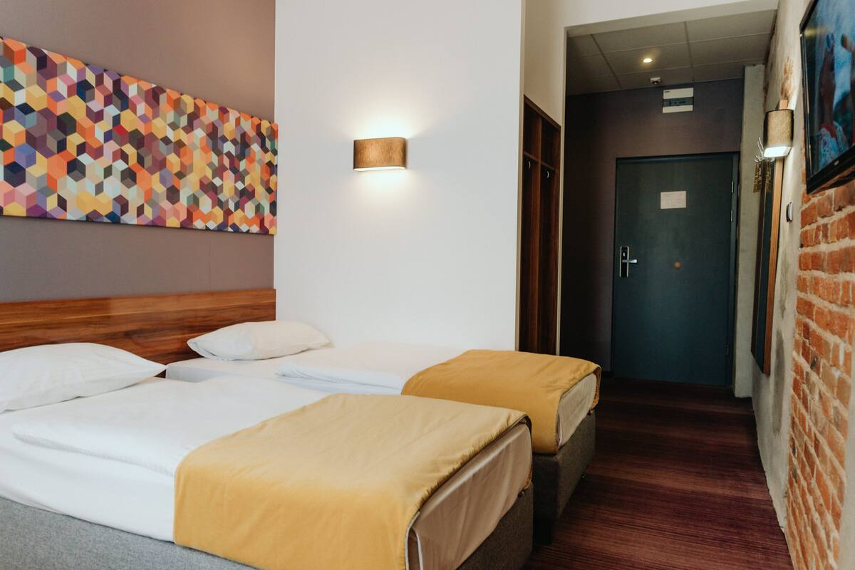 Habitación de hotel con dos camas, cabecero de madera, cuadro de pared con arte geométrico de colores, lámpara de pared y pared de ladrillo con un televisor.