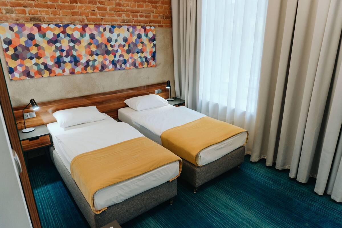 Habitación de hotel con dos camas, una pared de ladrillo, un cuadro abstracto colorido, una mesita de noche con lámpara y una ventana con cortinas.
