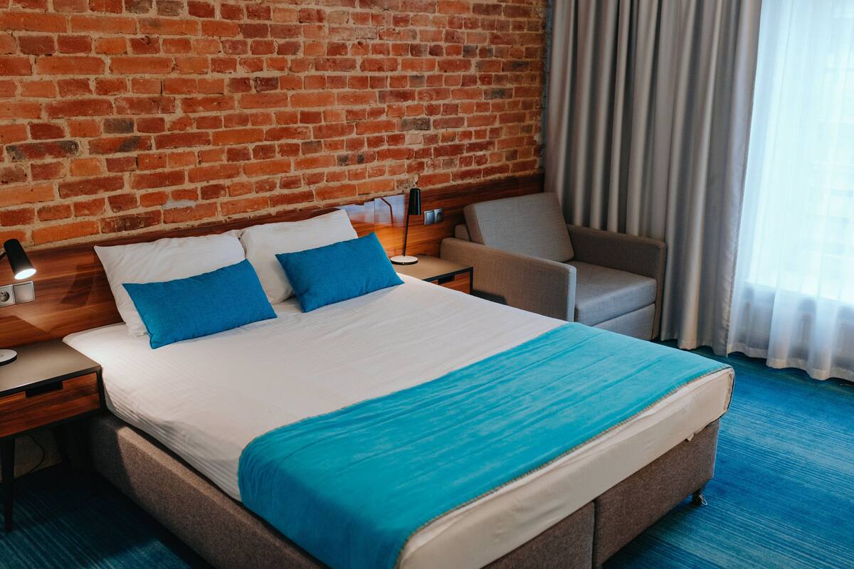 Habitación de hotel con pared de ladrillo, cama con ropa de cama azul y blanca, una silla, una mesita de noche con lámpara y una ventana con cortinas.