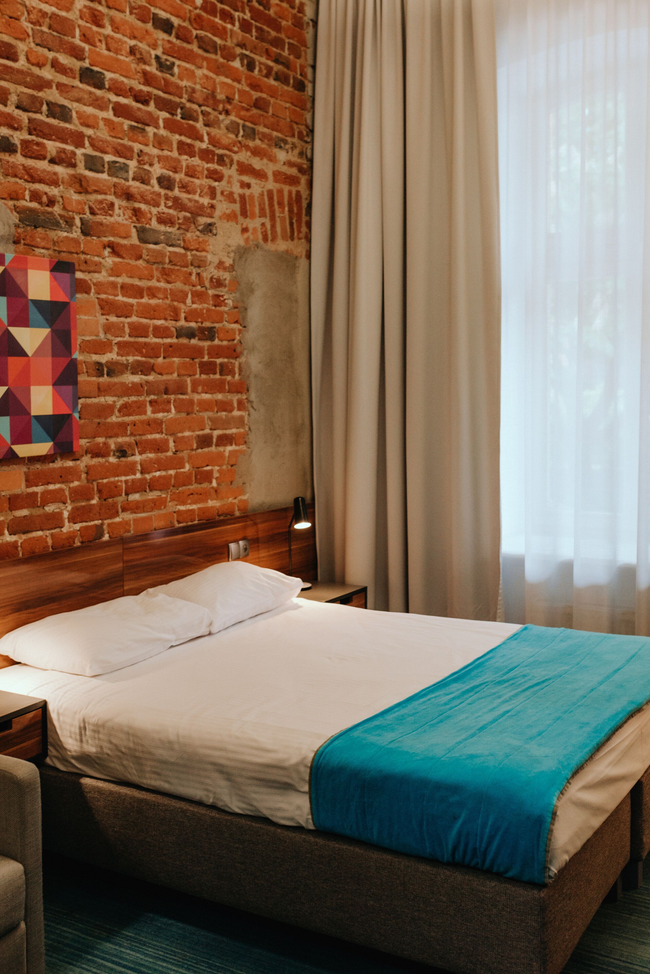 A hotel room with a brick wall, a bed with white linens and a blue blanket, a wooden headboard, a nightstand, and a lamp.