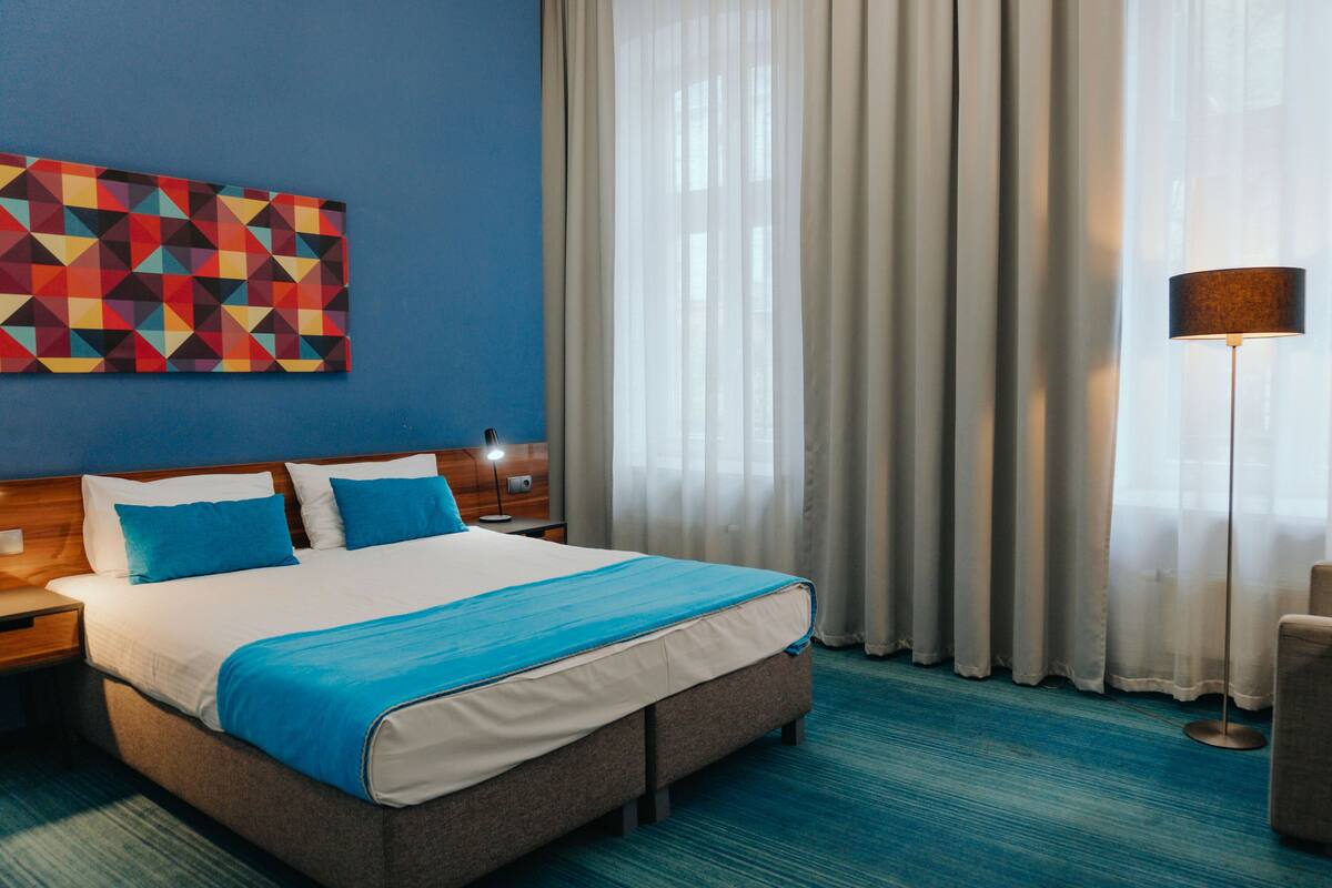 Habitación de hotel con una cama grande, ropa de cama azul y blanca, una obra de arte geométrica en la pared, una mesita de noche con una lámpara y vistas al exterior a través de cortinas translúcidas.