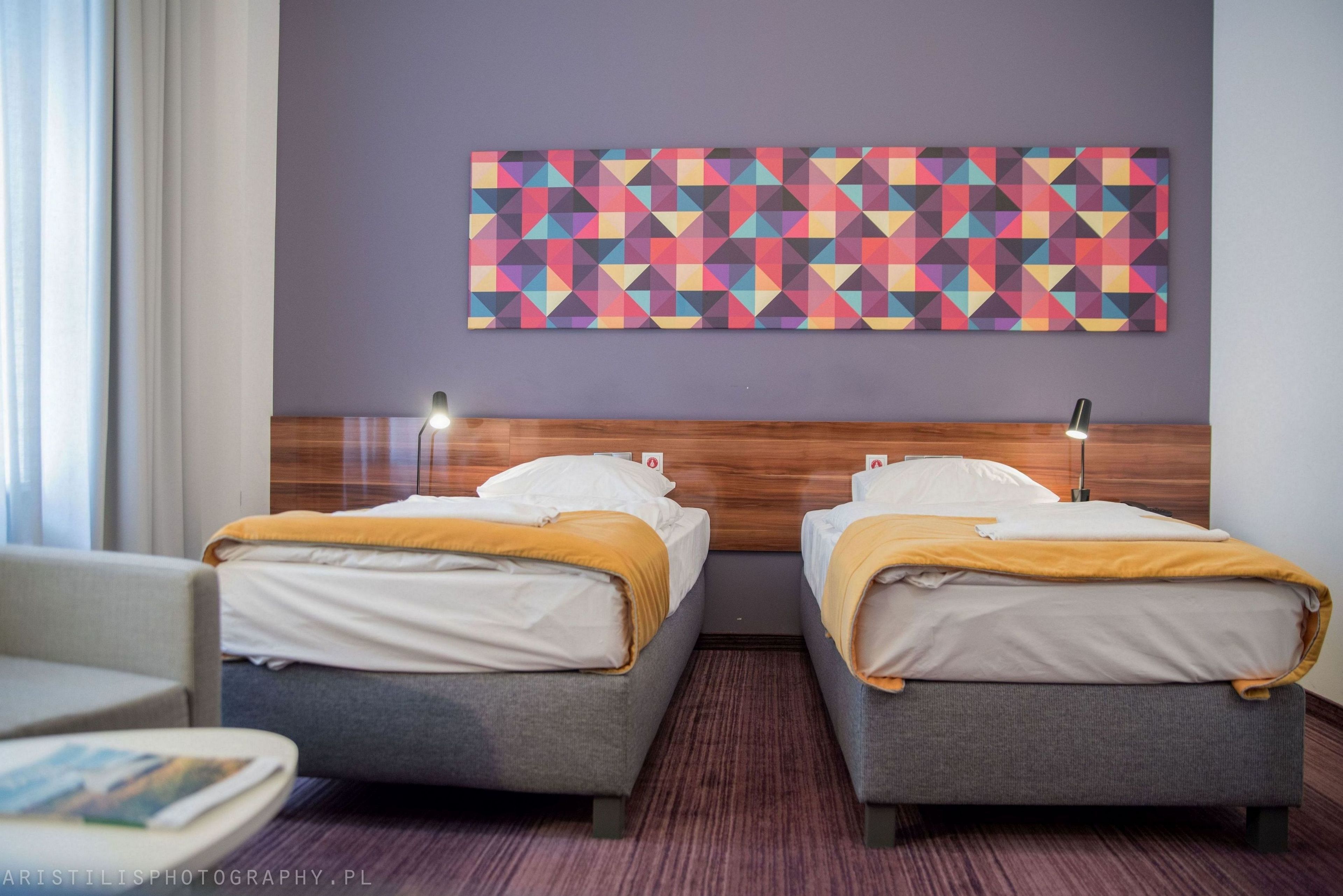 A hotel room with two beds, a wooden headboard, a colorful geometric wall art, and a small table with a magazine.