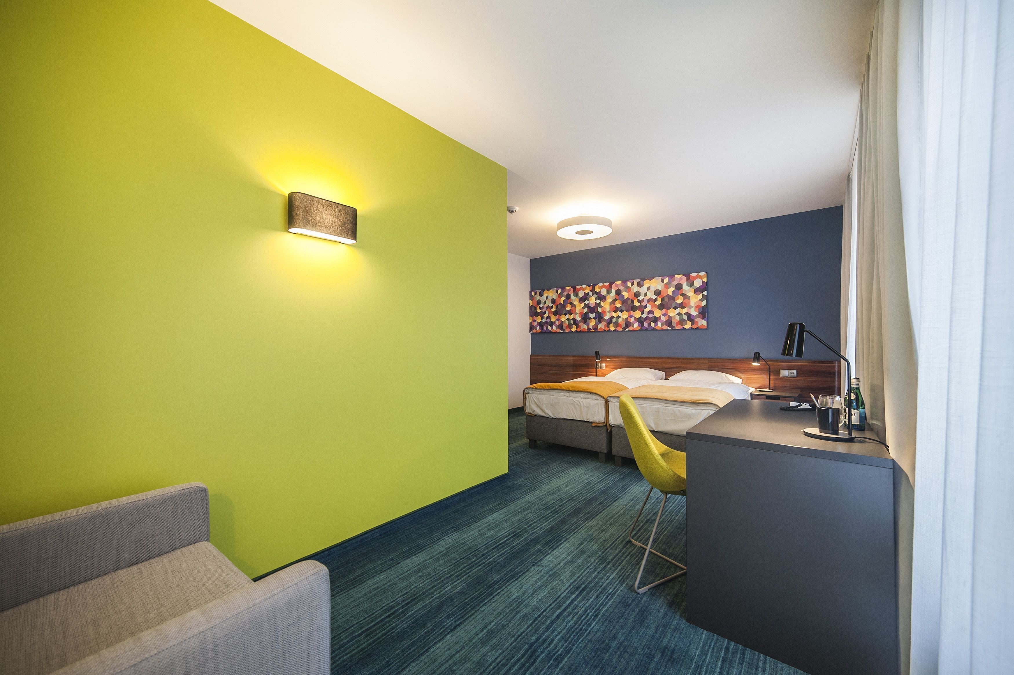 A hotel room with a bed, a desk, a chair, and a colorful abstract painting on the wall.