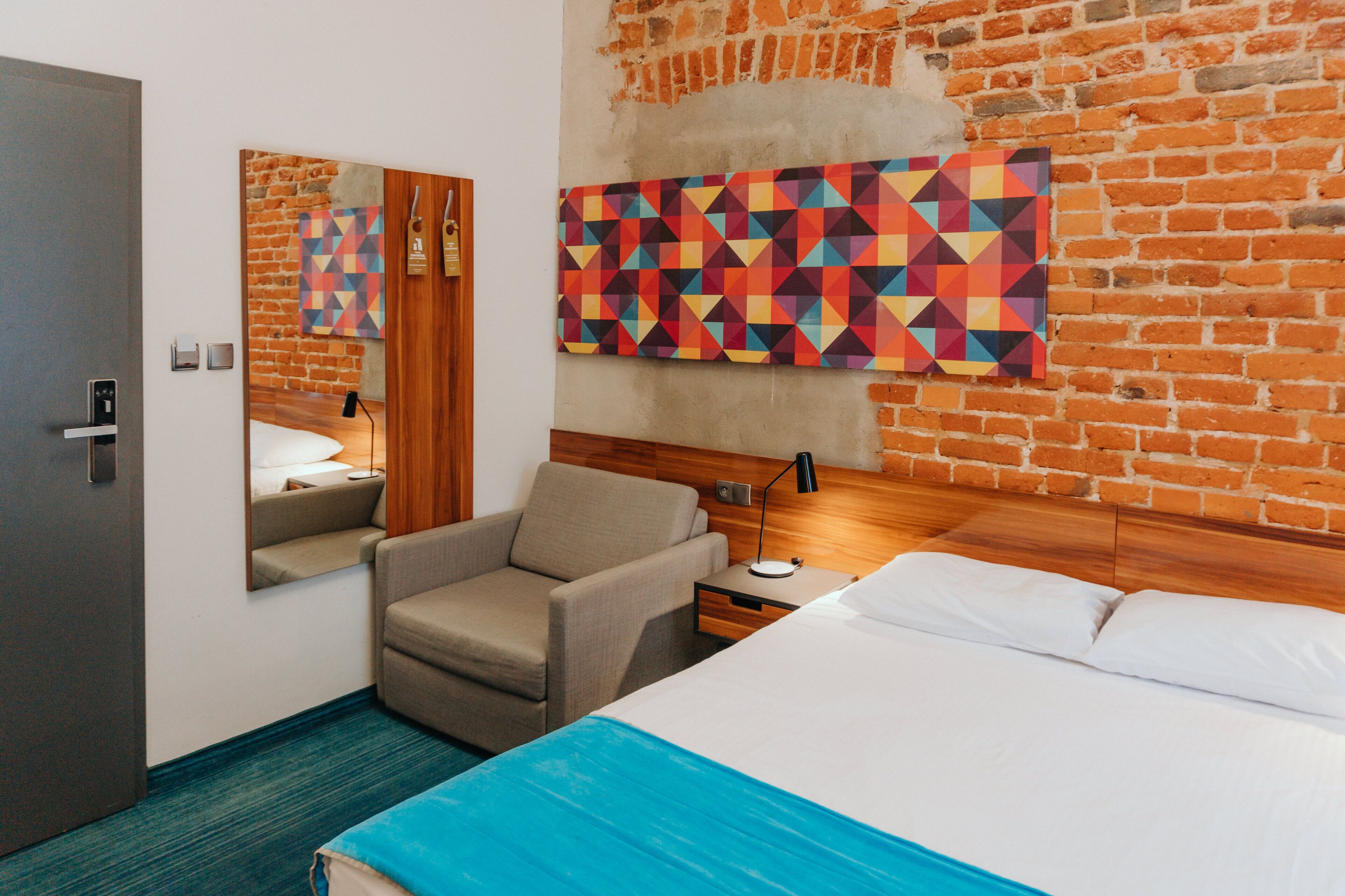 A hotel room with a brick wall, a bed, a chair, a nightstand, and a mirror.