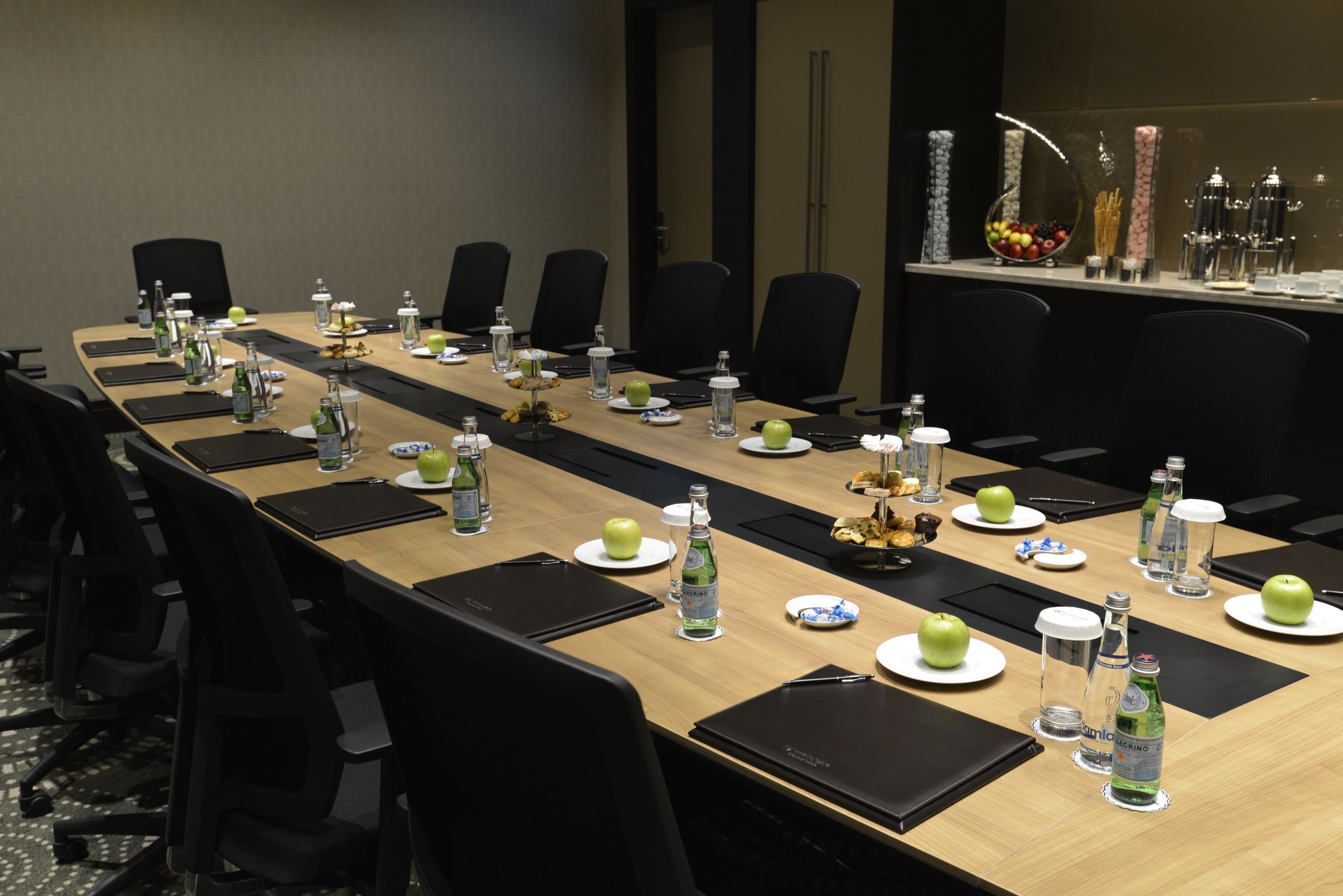 Un salón de conferencias con una larga mesa dispuesta para una reunión, equipado con botellas de agua, manzanas y un frutero.