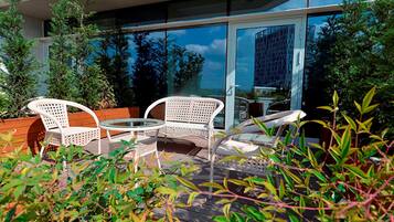 Un patio al aire libre con mobiliario de mimbre blanco, una mesa de vidrio y vista a un edificio moderno a través de grandes puertas de cristal.