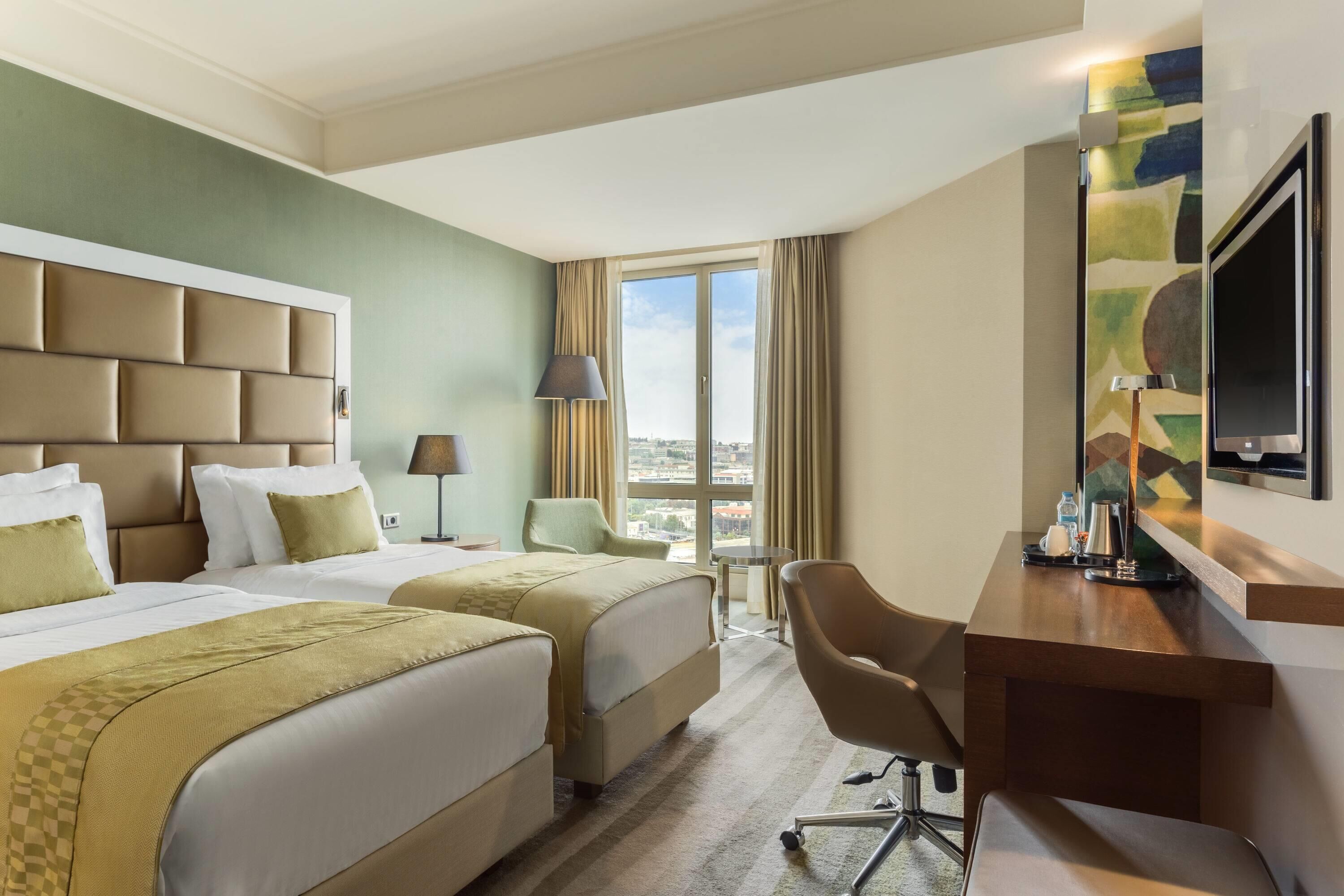 A hotel room with two beds, a desk, a chair, a TV, and a window with a city view.