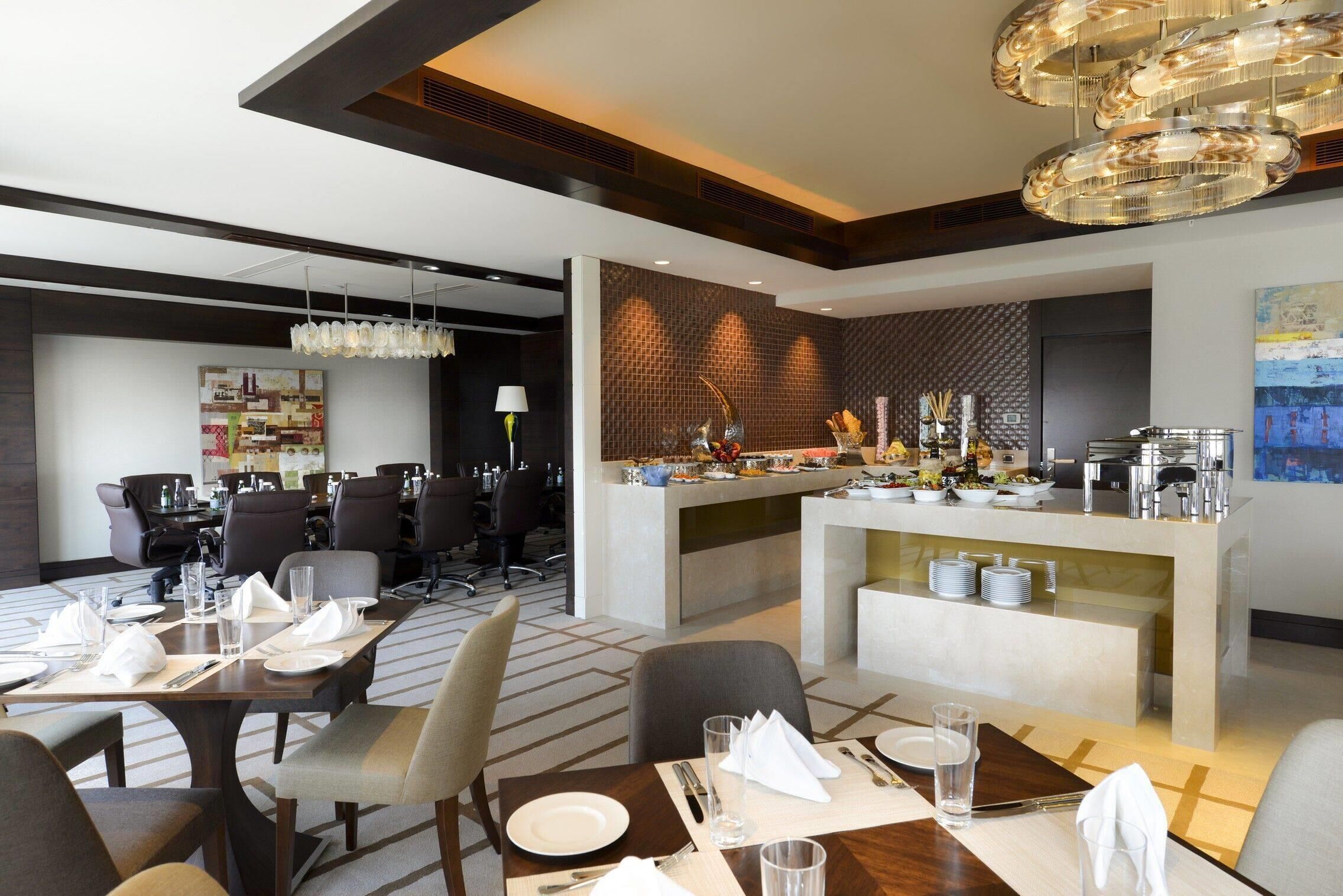 Un comedor de hotel moderno con una barra central, varias mesas dispuestas para comer y una lámpara contemporánea.