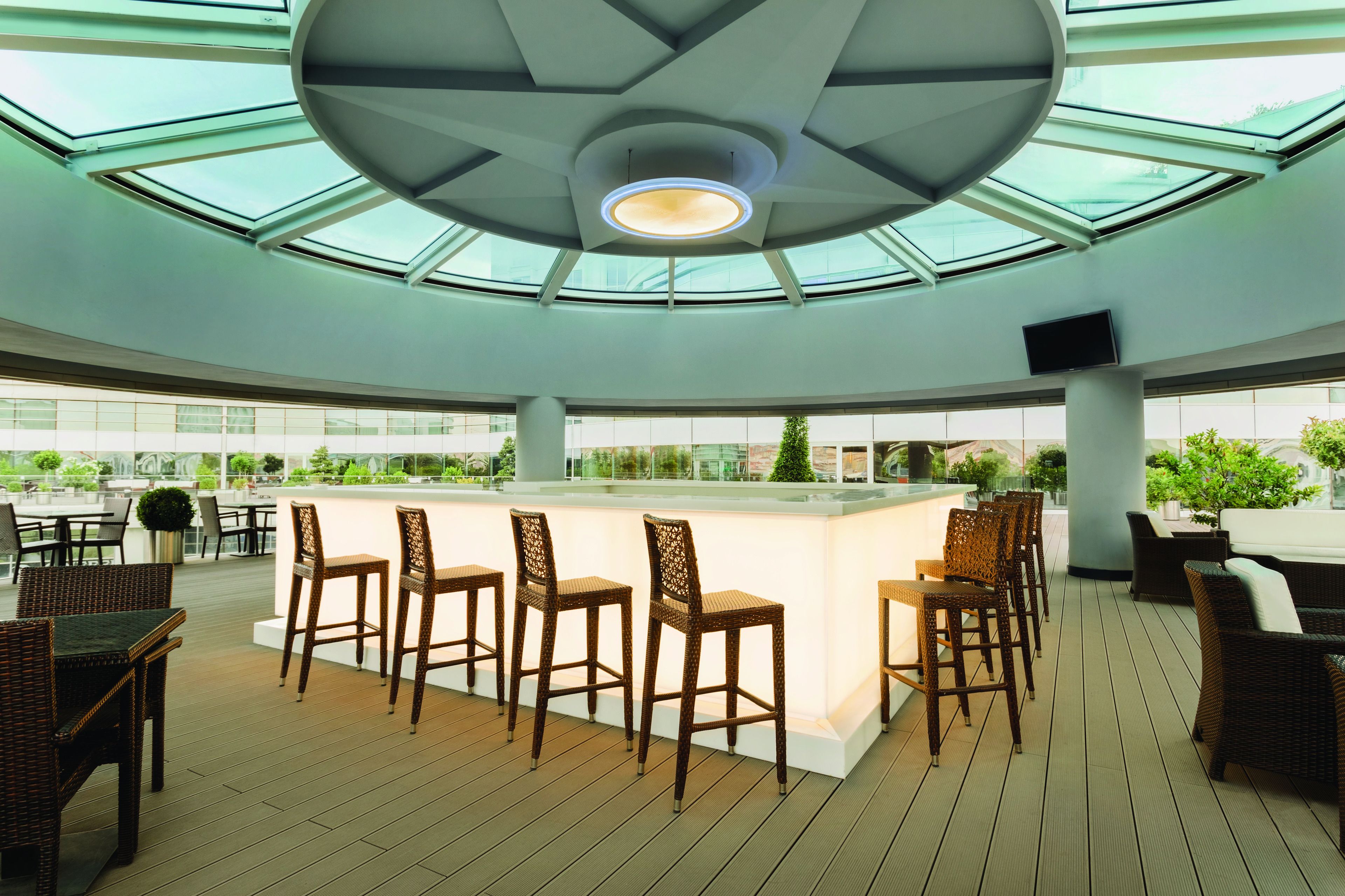 A spacious outdoor bar area with a central island, surrounded by wicker chairs and tables, under a large glass ceiling.