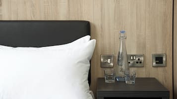 A neatly made bed with white linens and a black headboard, a bedside table with a water bottle and glasses, and a wall-mounted light switch.