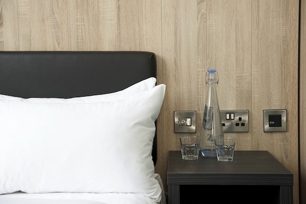 A neatly made bed with white linens and a black headboard, a bedside table with a water bottle and glasses, and a wall-mounted light switch.