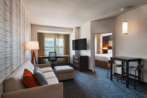 A hotel room with a sofa, a desk with a computer, a TV, and a small dining area.
