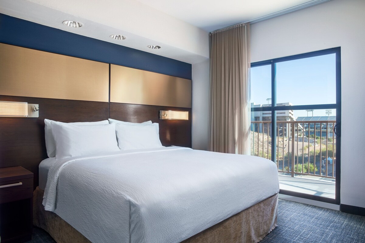 A hotel room with a large bed, a wooden headboard, a nightstand, and a view of the outside through a large window.