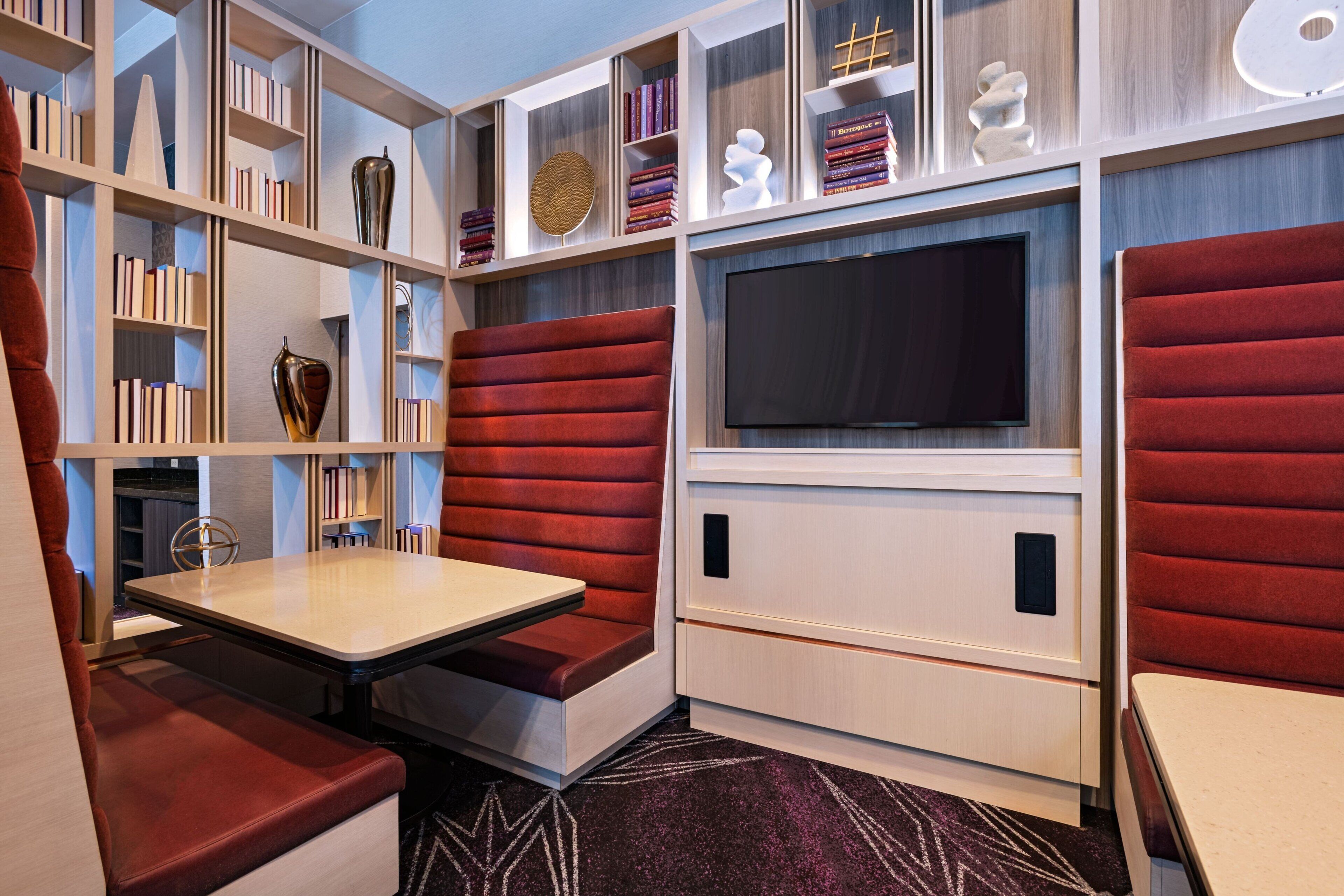 Un salon moderne doté d’un téléviseur encastré, de sièges rembourrés en tissu rouge et d’une bibliothèque remplie de livres et d’objets décoratifs.